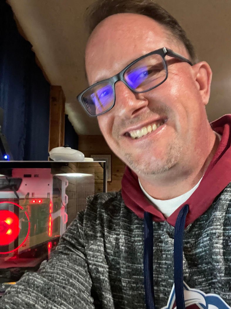 A man wearing glasses and a hoodie smiles at the camera, with a lit computer case featuring red lights visible in the background.