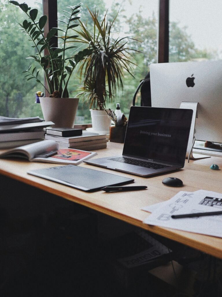 A wooden desk with a laptop, desktop computer, notebooks, papers, potted plants, and office supplies near a large window overlooking greenery.