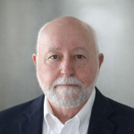 An older man with a white beard and bald head, wearing a dark blazer and white shirt, poses against a plain, light gray background.
