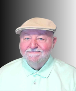 An older man with a white beard and mustache wears a light-colored flat cap and mint green collared shirt, smiling in front of a black-to-white gradient background.
